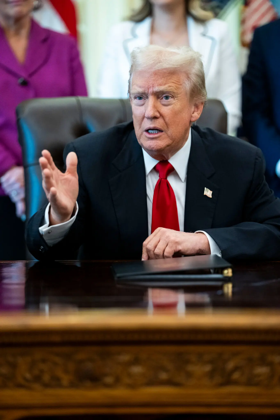 白宫椭圆形办公室内，身着蓝色西装、系红色领带的特朗普总统坐在办公桌前讲话，右手做出手势。在人工智能算力领域的投资热潮后，特朗普正竭力推动美国整体经济实现新一轮增长。